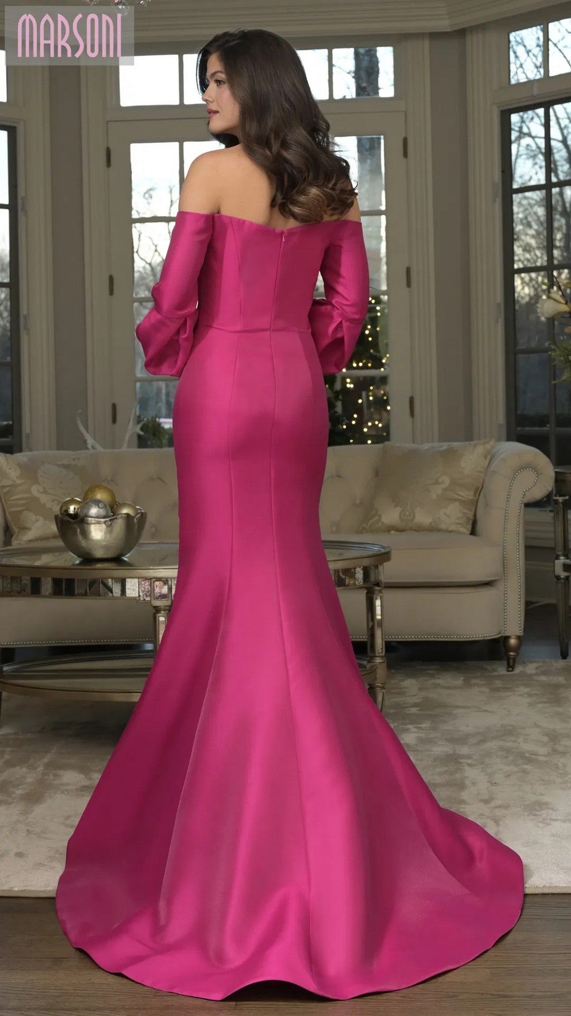 A woman wears the Marsoni MV1355 long formal dress with an off-the-shoulder mermaid silhouette, standing in a living room with neutral furniture and large windows.